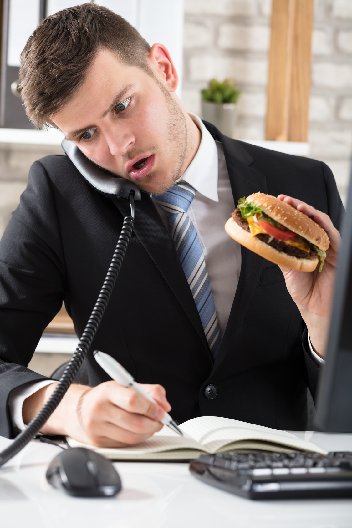 ein Mitarbeiter im Büro telefoniert, macht Notizen und isst zeitgleich einen Burger.